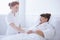 Senior grey woman lying in white hospital bed with young helpful nurse holding her hand