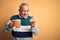 Senior handsome man drinking jar of beer standing over isolated yellow background Beckoning come here gesture with hand inviting