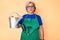 Senior hispanic grey- haired woman wearing gardener apron and gloves holding watering can thinking attitude and sober expression