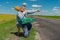 Senior hitch-hiker having short rest on a rural roadside with an ancient green suitcase and mandolin