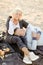 Senior husband lying on wife knees and plaid, champagne and fruits on sand beach.