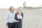 Senior husband walking on sand beach with blonde wife talking by smartphone.