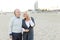 Senior husband walking on sand beach with blonde wife talking by smartphone.