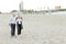 Senior husband walking on sand beach with blonde wife talking by smartphone.