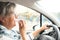 A senior lady with gray hair and eyeglasses driving a car. Interior. Side view. Lip gloss in the hand