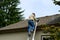 Senior man climbing an aluminum extension ladder to a home rooftop with a container of moss killer granules