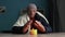 Senior man dressed in a warm winter hat and a scarf sits at home at a table and warms his hands from a burning candle