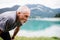 A senior man with earphones standing by lake in nature, doing exercise.