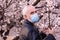 Senior man in face mask, standing in a flowering apricot blossom garden, breathing of spring flowers during pandemic. Pensioner wa