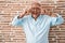 Senior man with grey hair standing over bricks wall doing peace symbol with fingers over face, smiling cheerful showing victory