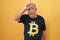 Senior man with grey hair wearing bitcoin t shirt making fun of people with fingers on forehead doing loser gesture mocking and
