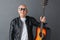 Senior man in leather jacket and sunglasses standing isolated on gray holding guitar looking camera positive