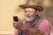 Senior man in straw hat listening music from cellular over headphones while sitting outdoor against clay wall