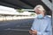 A senior man wearing medical mask due to coronavirus standing under the metal structure of a deserted parking looking at his smart