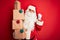 Senior man wearing Santa Claus costume holding tower of gifts over isolated red background screaming proud and celebrating victory