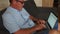 Senior man working on laptop at home. He wears a blue shirt and wears glasses for sight. Remote employment, freelancing