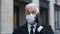 Senior masked businessman with gray hair in black coat, gloves speaks on phone.