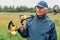 Senior numismatist wearing cap and jacket holding metal detector over his shoulder and looking away, treasure hunter searching