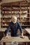 Senior potter standing and leaning on table against shelves with pottery goods at workshop