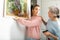 Senior woman with adult daughter hanging picture on wall at home