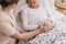 Senior woman lying in bed, helpful doctor in beige uniform supporting her