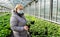 Senior woman in a medical mask chooses a geranium sprout from a variety of bright green seedlings in flower pots in a greenhouse