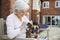 Senior Woman Sitting On Bench With Pet French Bulldog In Assisted Living Facility