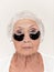 Senior woman at studio isolated on white wall looking at camera and making masks under eyes