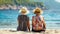 Senior women in straw hats sitting on beach, rear view, summer travel outdoor concept