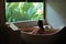 Sensitive young woman enjoying in spa. Luxury stone bath tub with jungle view near window. Natural organic tropical petals in the