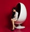 Sensual melancholic lady sexy brunette in white long jacket and high-heeled shoes sitting posing in a modern oval chair on red