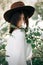 Sensual portrait of beautiful hipster woman in hat smelling  white flowers in spring. Stylish calm boho girl posing in blooming