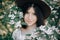 Sensual portrait of beautiful hipster woman in hat smiling in white spring flowers. Stylish calm boho girl posing in blooming tree