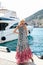 Sensual woman with blond hair in luxurious dress with accessories posing near yacht in dock