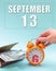 September 13th. Hand holding an orange alarm clock, a wallet with cash and a calendar date. Day 13 of month.