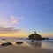 Serene beachscape at twilight with orange-hued sky, small islet crowned with foliage stands amid tranquil tide