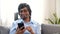 Serene smiling young indian man sitting down on the sofa at home with a smartphone
