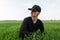 Serious attractive man in a black vintage shirt in a fashionable baseball cap sits in the green grass in a field.