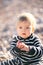 Serious baby in striped overalls sitting on a pebble beach holding folded palms in front of him