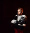 Serious blond boy in hockey uniform with hockey skates and helmet standing and looking aside over dark background