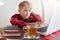 Serious blond little boy in red shirt sitting in front of open laptop, staring at screen with concentrated look drinking tea and s