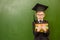 Serious boy in graduation cap with books standing near green chalkboard