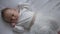 Serious Caucasian infant lying on white bed looking away. Top view portrait of charming cute curios baby girl in white
