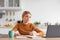 Serious caucasian teenage girl blond sits at table, looks at laptop, studies in minimalist kitchen