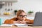 Serious caucasian teenage girl blonde in glasses sits at table with laptop, watches video lesson in minimalist kitchen