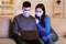 Serious couple in face mask using laptop sitting in sofa