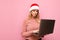 Serious cute girl in santa hat stands on a pink background with a laptop in her hands and looks focused. camera. Lady freelancer