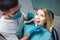 Serious dentist in mask and white robe stand beside client and checking teeth condition. Young woman sit in chair and