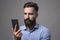 Serious dissatisfied young bearded man holding and watching smartphone