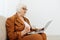 a serious, experienced, elderly woman is sitting working from home on the couch with a laptop on her lap and menacingly